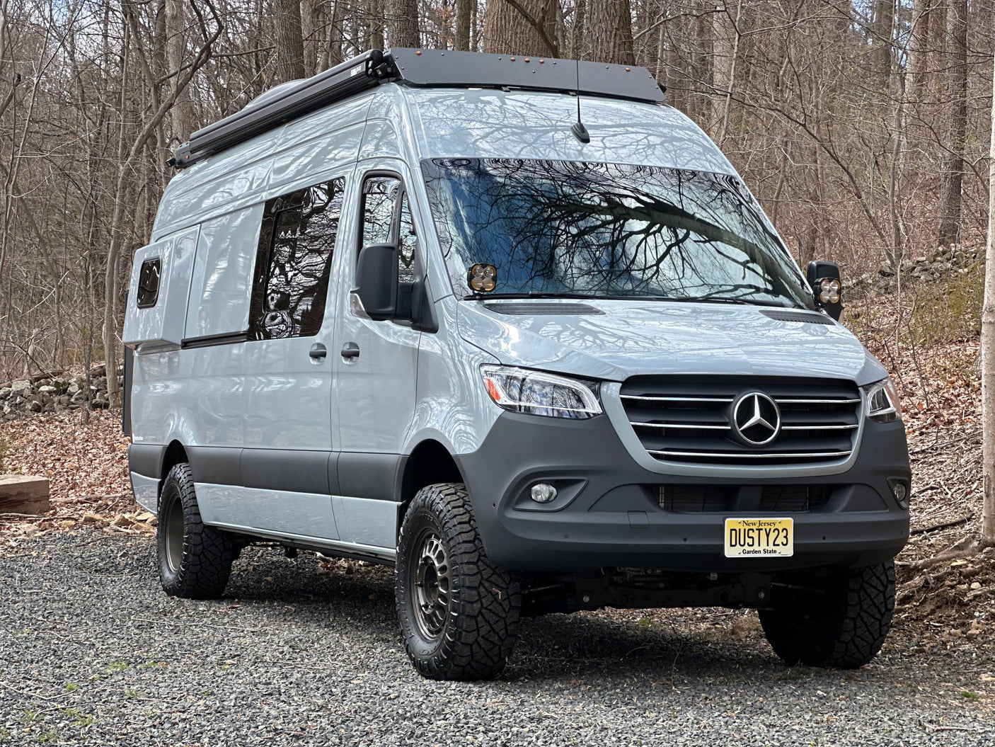 Light grey, lifted Mercedes-Benz Sprinter custom campervan with off-road tires, parked in woods. Ideal RV rental.