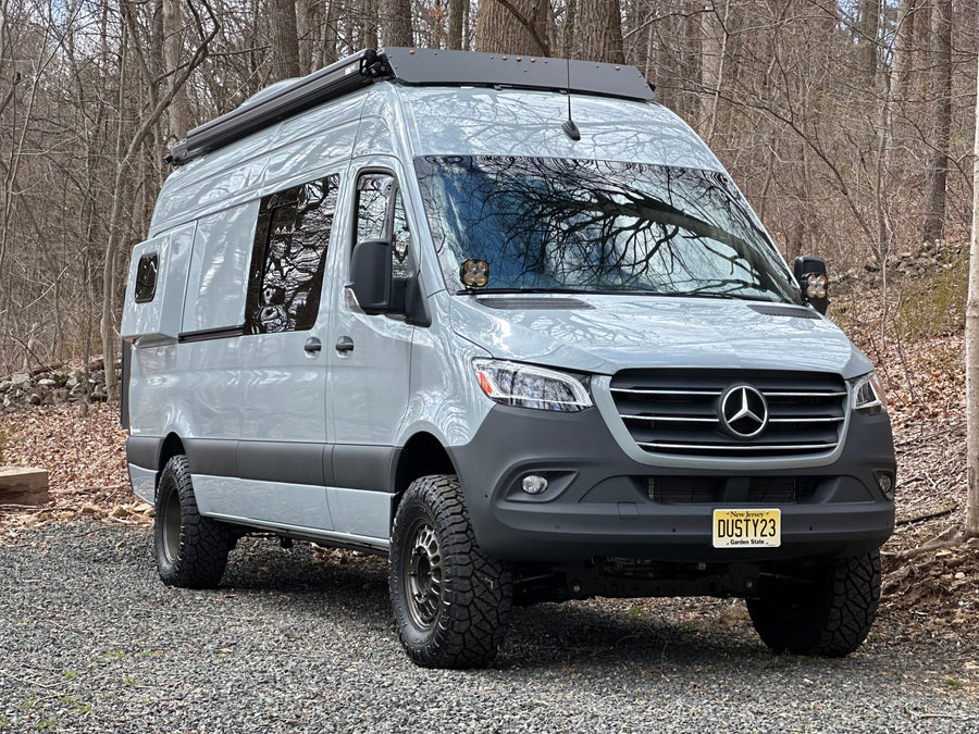 Light grey, lifted Mercedes-Benz Sprinter custom campervan with off-road tires, parked in woods. Ideal RV rental.