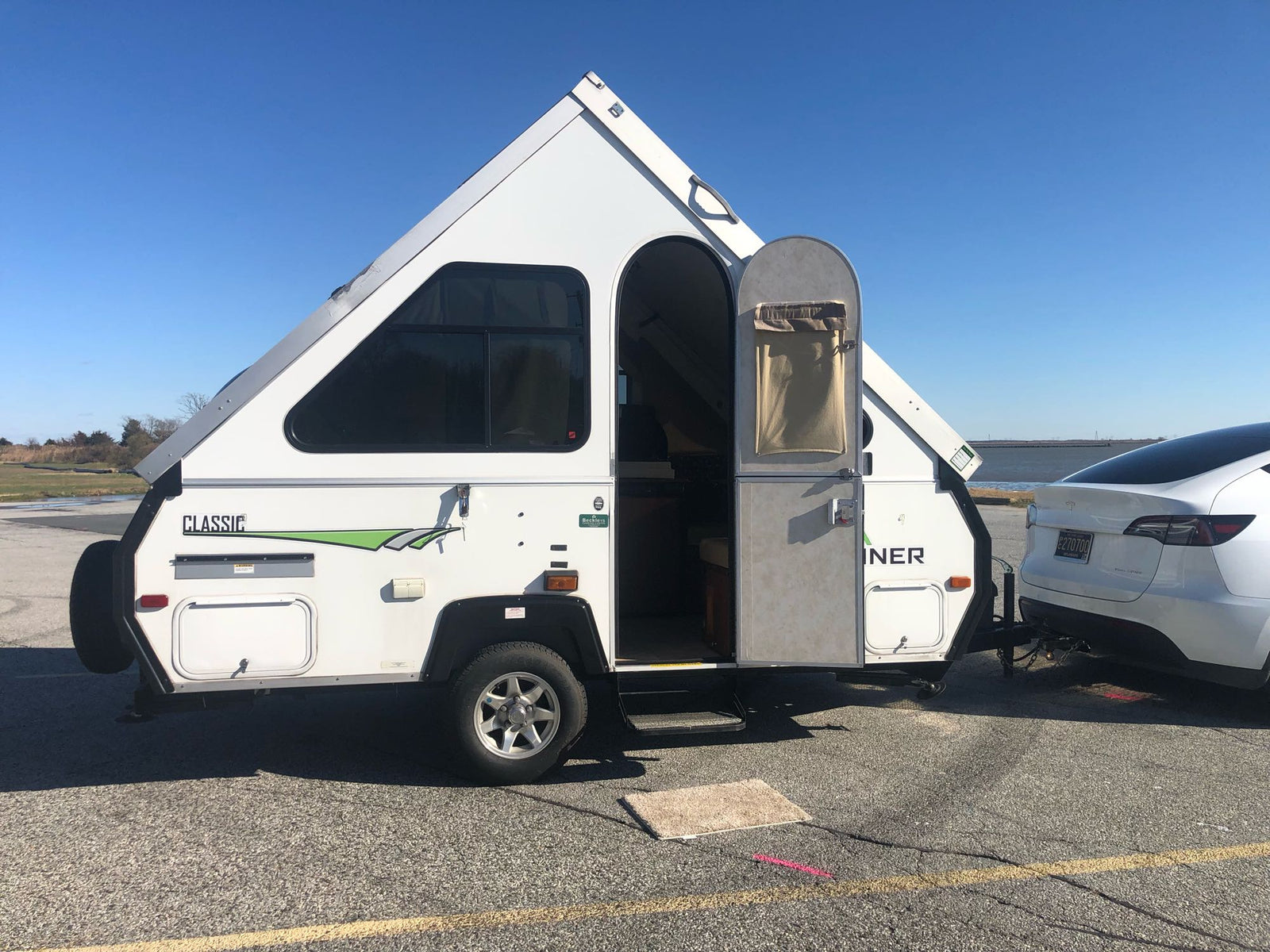 sits on asphalt under a clear sky.|White Tesla Model Y towing an A-liner RV