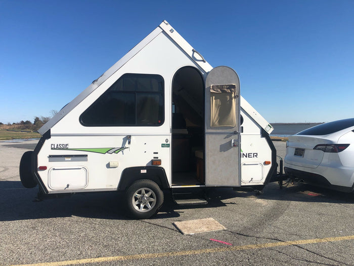 sits on asphalt under a clear sky.|White Tesla Model Y towing an A-liner RV