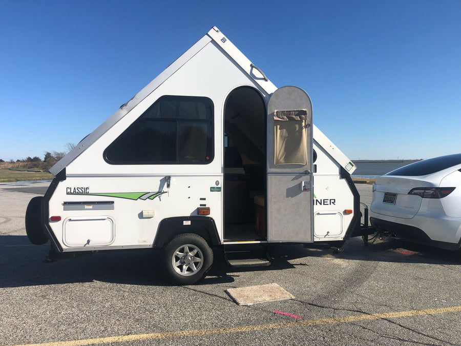 sits on asphalt under a clear sky.|White Tesla Model Y towing an A-liner RV