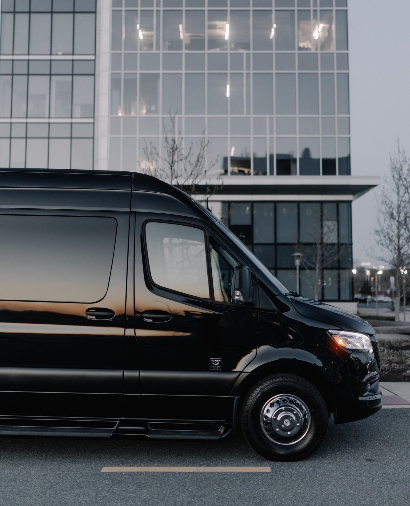 A polished black luxury van, suitable for an RV rental, sits on a street with a large, modern glass building behind it.