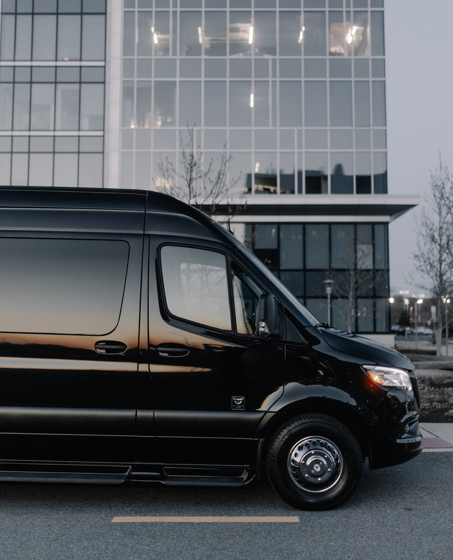A polished black luxury van, suitable for an RV rental, sits on a street with a large, modern glass building behind it.