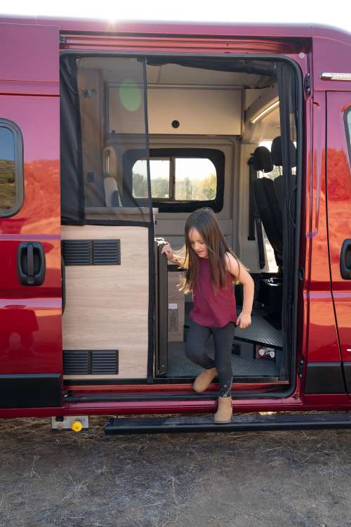 woman on laptop at kitchen counter. Functional camper van for mobile living.|View of a clean RV rental camper van interior with built-in bed