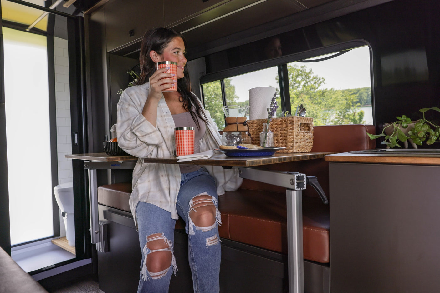 Woman sips coffee in a modern rv rental, enjoying a scenic view of trees and water from her table in the camper's sleek interior.