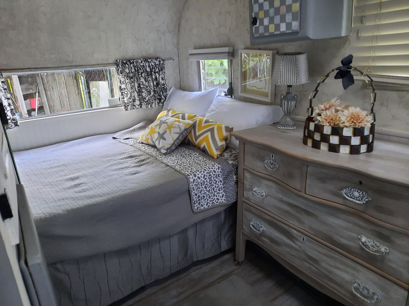 Inside a comfortable RV rental: a bedroom area with a grey bed, patterned pillows, distressed wood dresser, and unique lamps.