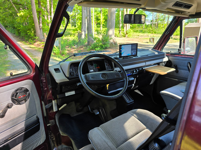 is parked on a gravel path by a serene lake with docks and lush trees.|Relaxing by the river: a maroon camper van