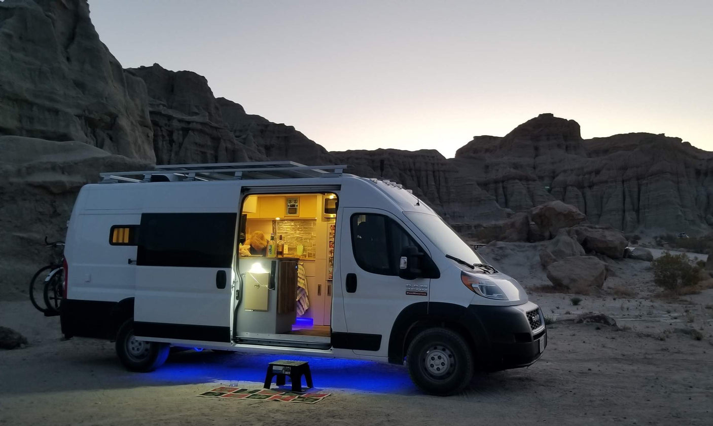 RV rental camper van with open illuminated kitchen, blue underglow, & bike rack, parked in a scenic desert landscape.