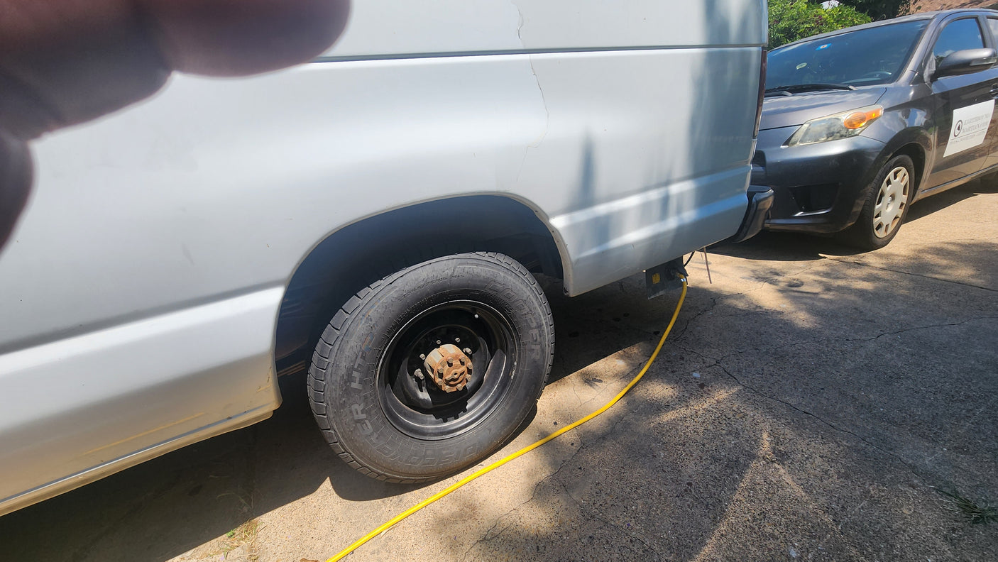 RV rental white van: Maintenance in progress. Rear wheel removed, exposing rusty hub. Yellow cable connected.