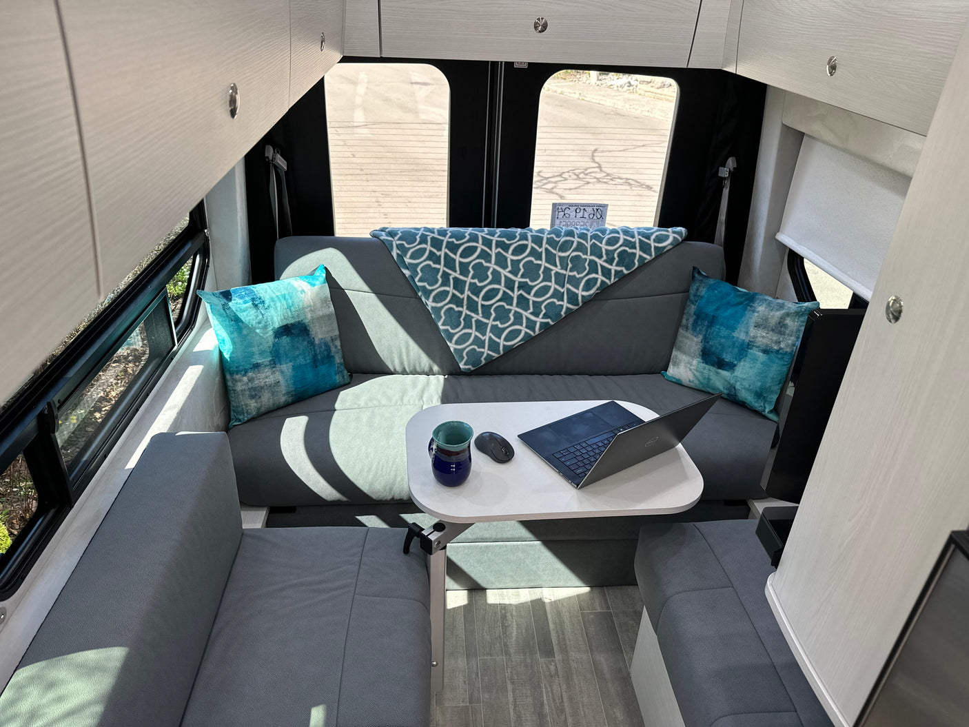 Cozy RV rental interior with grey seating, a white table with a laptop, and overhead cabinets, ideal for a mobile workspace.