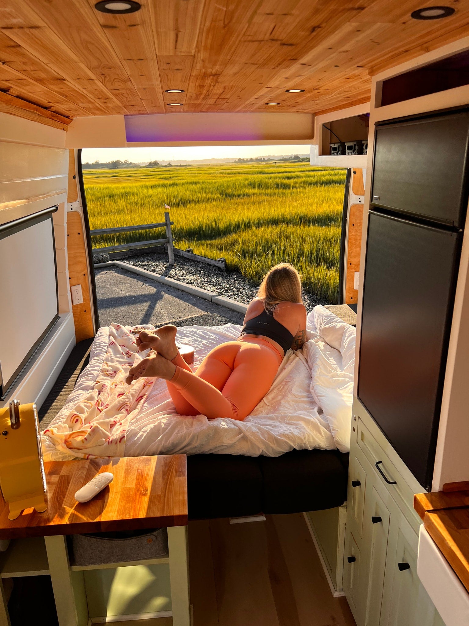 A person relaxes in an RV rental, looking out at a stunning sunlit marsh. Features a cozy bed and wooden interior.