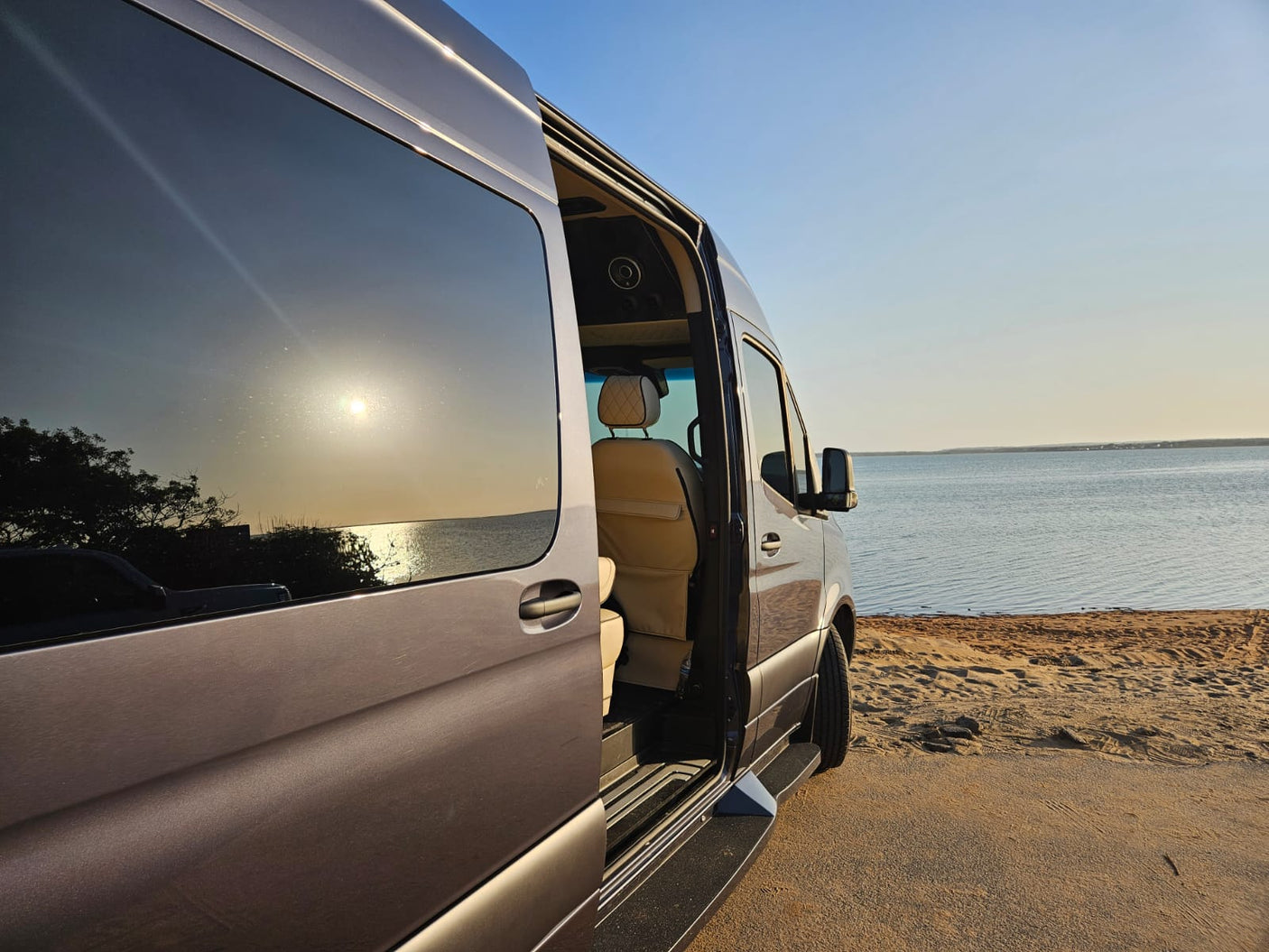 RV rental parked on a sandy shore by water. Open door reveals a comfortable interior at sunset.