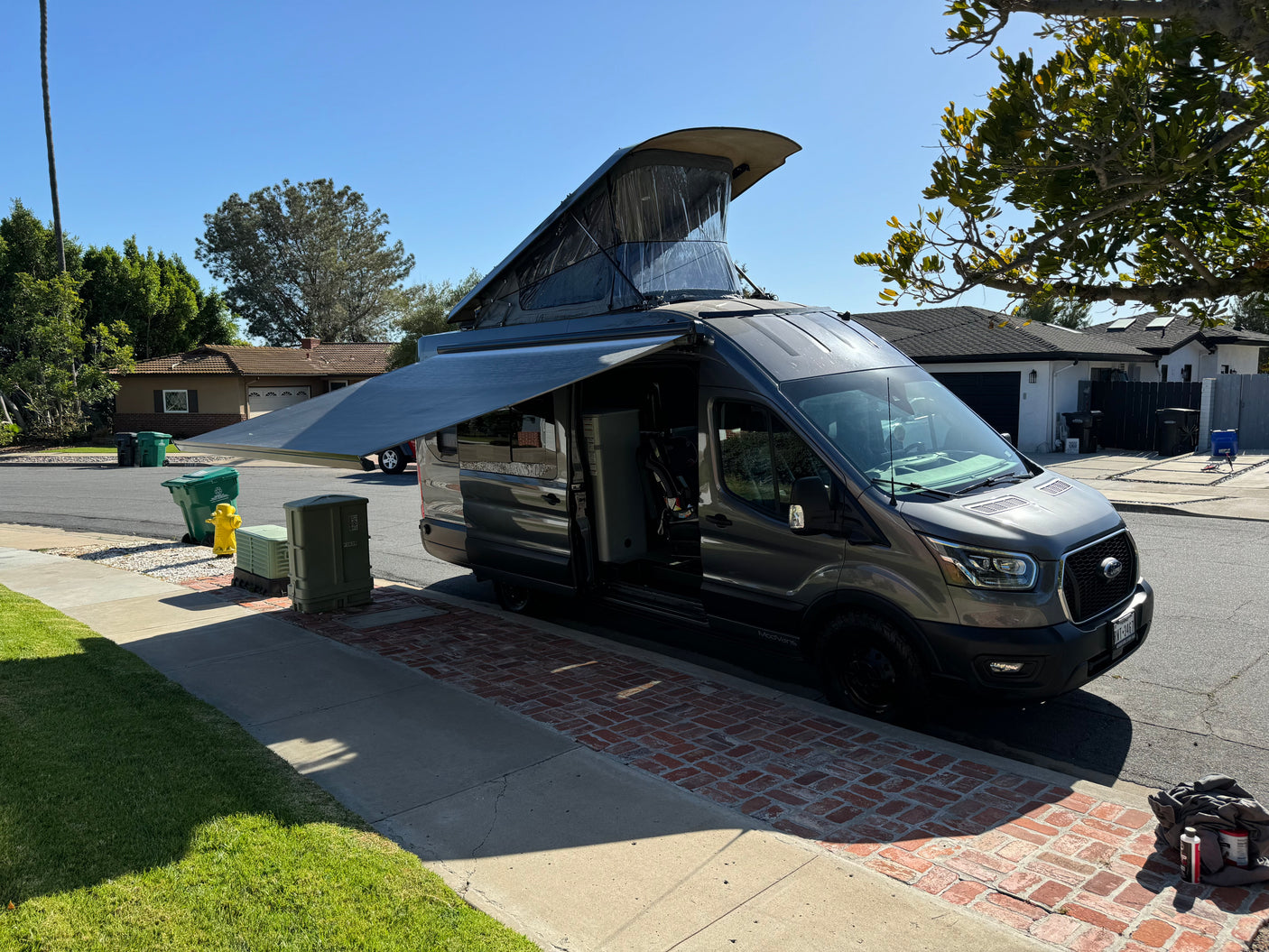 A gray ModVans camper van, an ideal rv rental, is parked with its pop-top roof and awning extended on a sunny day.