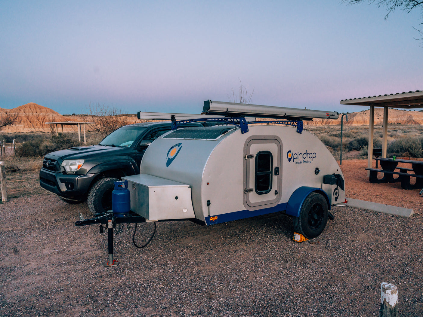 Teardrop RV rental camper and truck at a desert campsite, featuring hills and a picnic area.