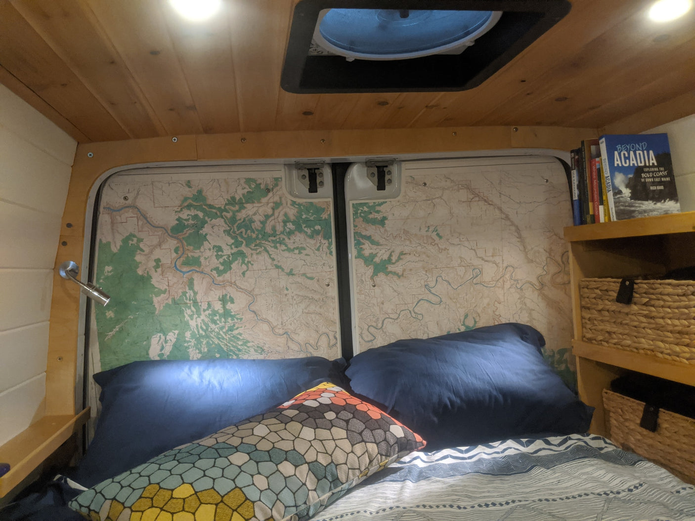 Cozy RV rental bedroom with maps on the back doors, a comfy bed, wooden ceiling, and travel books and storage baskets nearby.