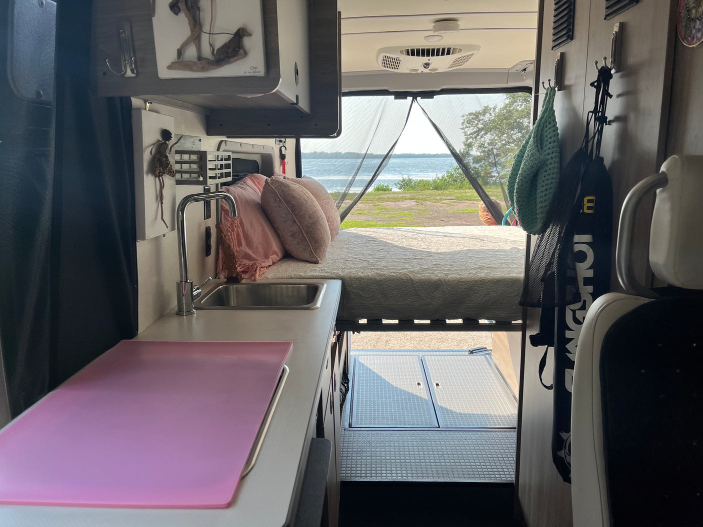 Inside an rv rental: a cozy bed at the rear overlooks a lake, with a kitchen counter & sink on the left. Ideal for travel.