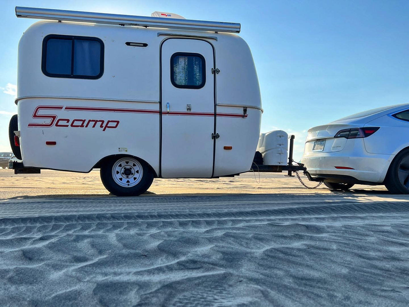 RV rental: A white Scamp travel trailer towed by a white Tesla on a sandy beach. Ideal for your next eco-camping trip!