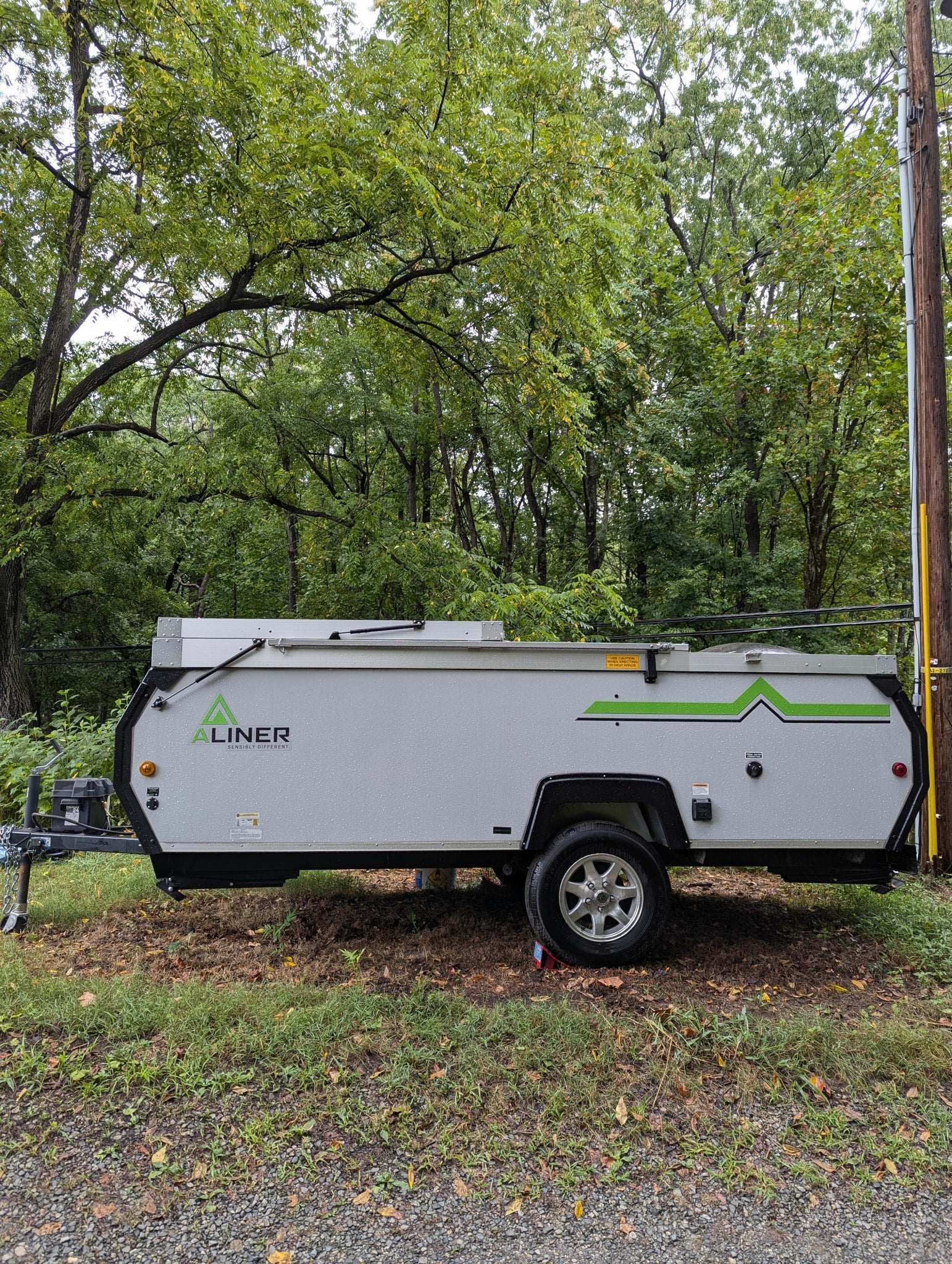 A light gray A-Liner pop-up RV rental with green accents, parked on gravel near lush trees. Perfect for compact camping adventures.