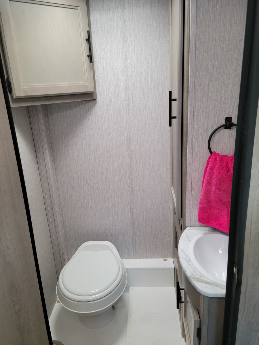 Compact RV rental bathroom with toilet, sink, and overhead storage. Features light grey walls and a bright pink towel.