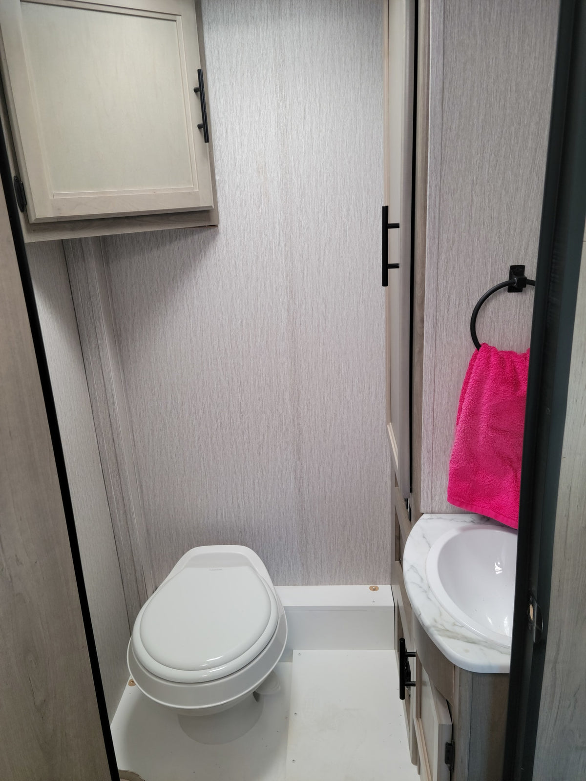 Compact RV rental bathroom with toilet, sink, and overhead storage. Features light grey walls and a bright pink towel. — photo 8