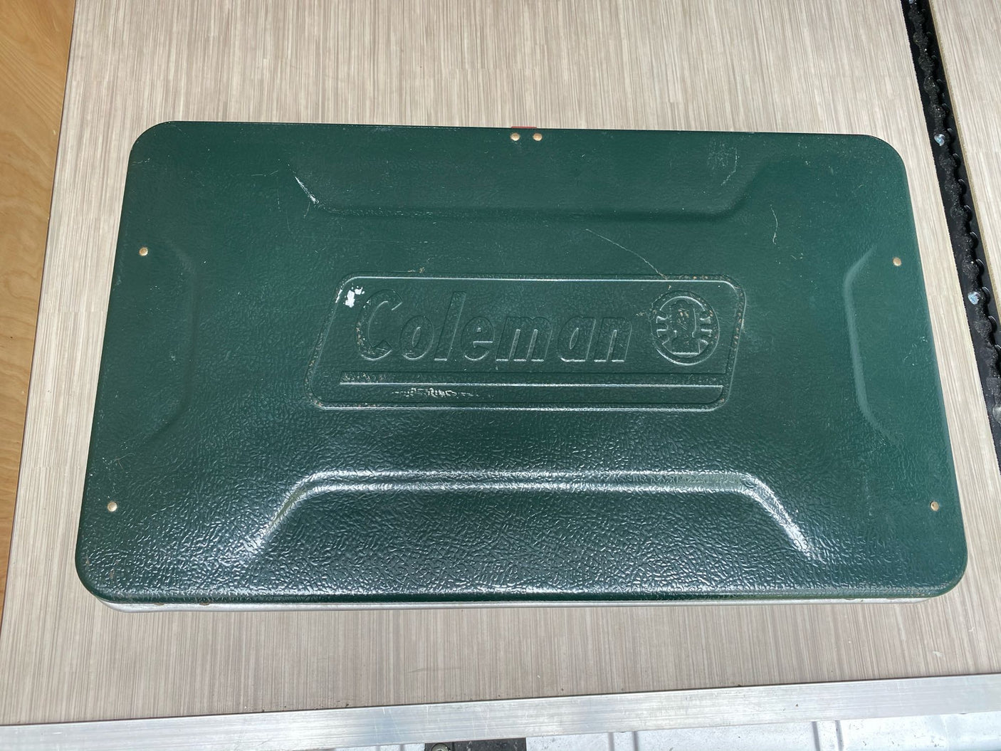 Dark green, textured Coleman camp stove (closed) on a light wooden surface. Ideal for your next RV rental adventure.