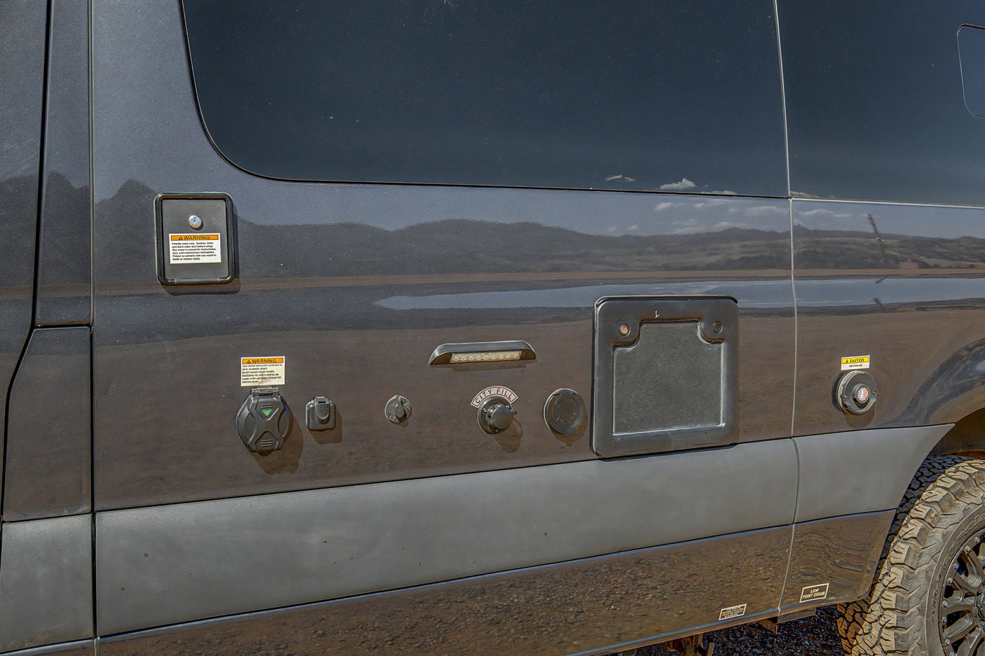 Dark grey RV rental camper van side panel with visible utility connections for fresh water, shore power, city fill, and air intake.