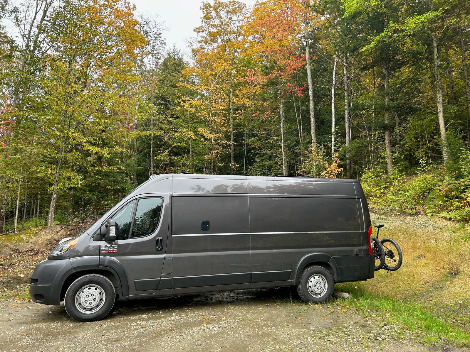 Grey camper van (RV rental) with bike rack, parked on dirt road amidst vibrant autumn forest. Ideal for outdoor adventures.
