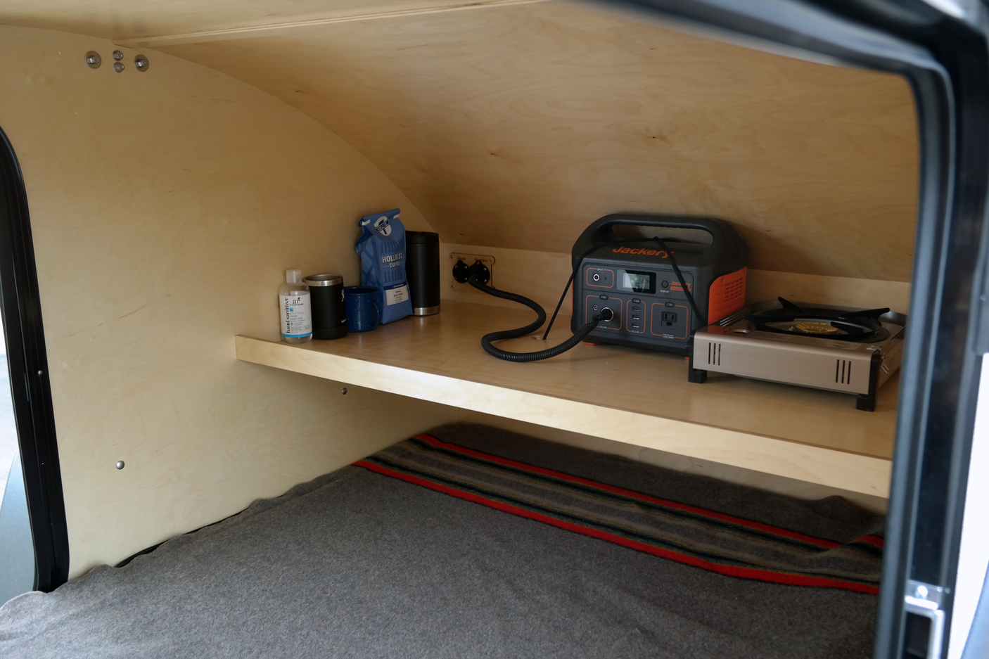 Inside a cozy RV rental: a shelf with a Jackery power station, portable stove, coffee, and bedding below.