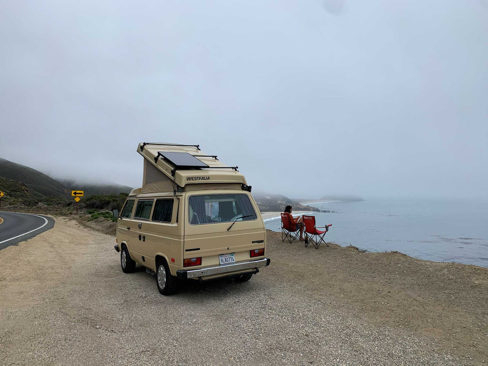Enjoy a coastal view from your RV rental. Tan VW Westfalia parked by ocean; person in chair watches misty coast.