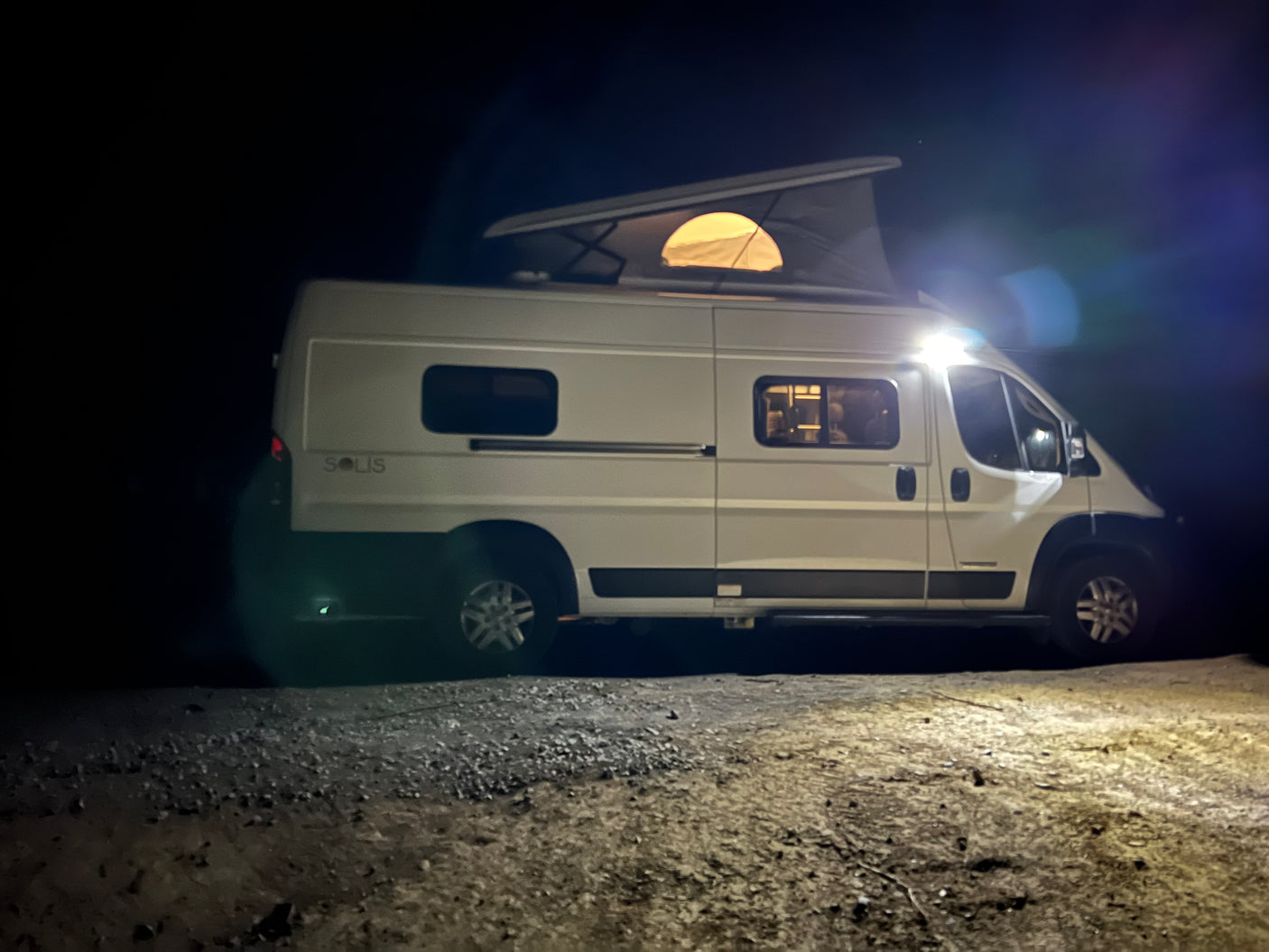 White RV rental camper van at night, with a lit pop-top tent and interior, parked on a gravel surface.