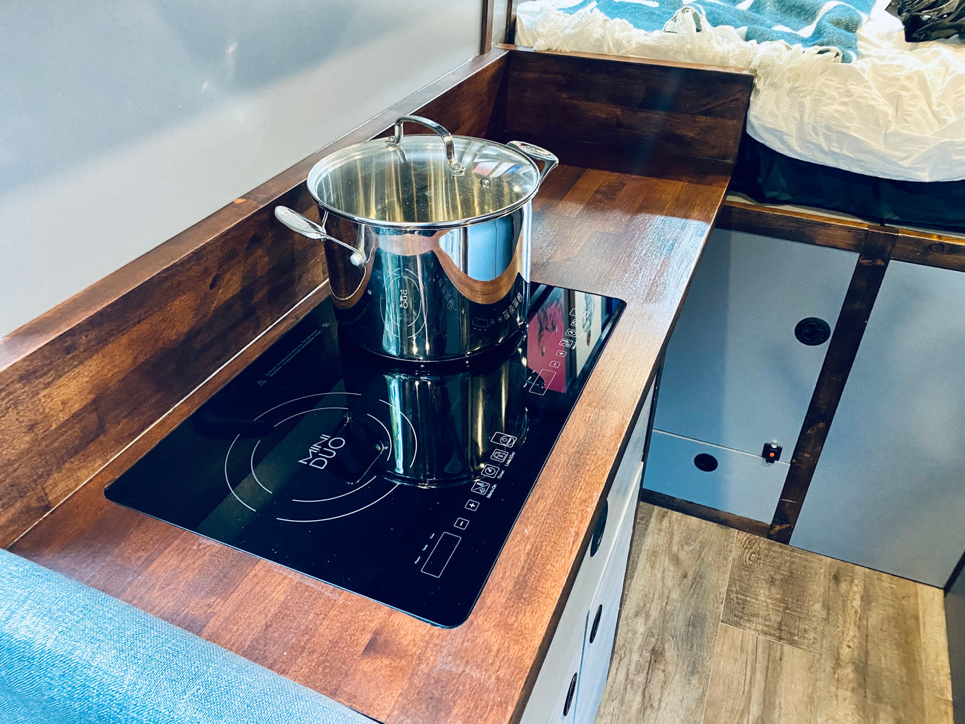 RV rental kitchen: compact setup with induction cooktop, pot on wooden counter, and adjacent sleeping area.