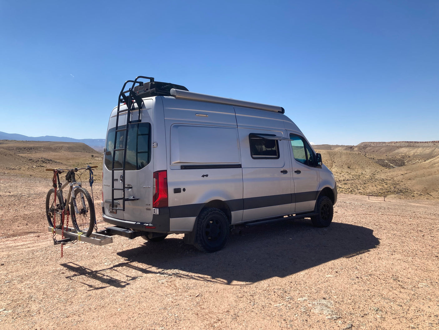 Silver RV rental van with mountain bikes on a rack, parked in a vast, sunny desert landscape.
