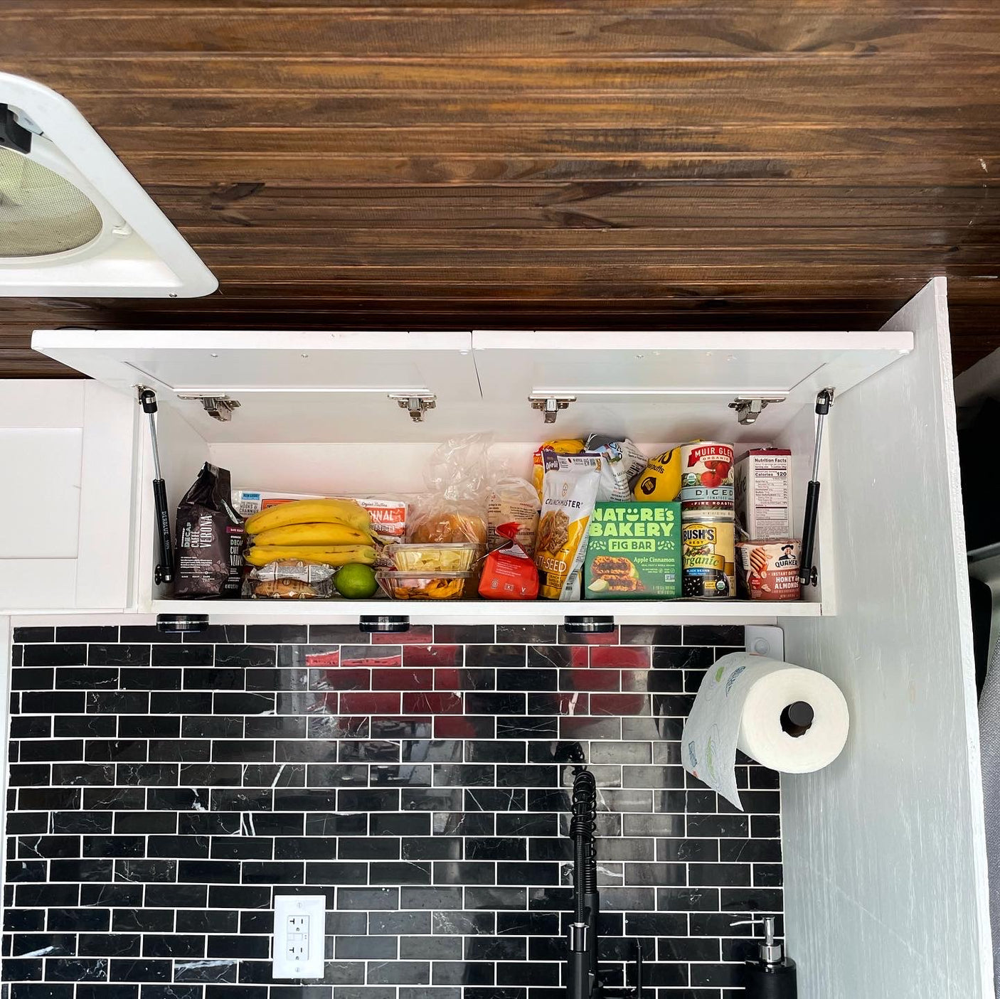 Compact RV rental kitchen features an open cabinet filled with food, black subway tile backsplash, and a paper towel holder.
