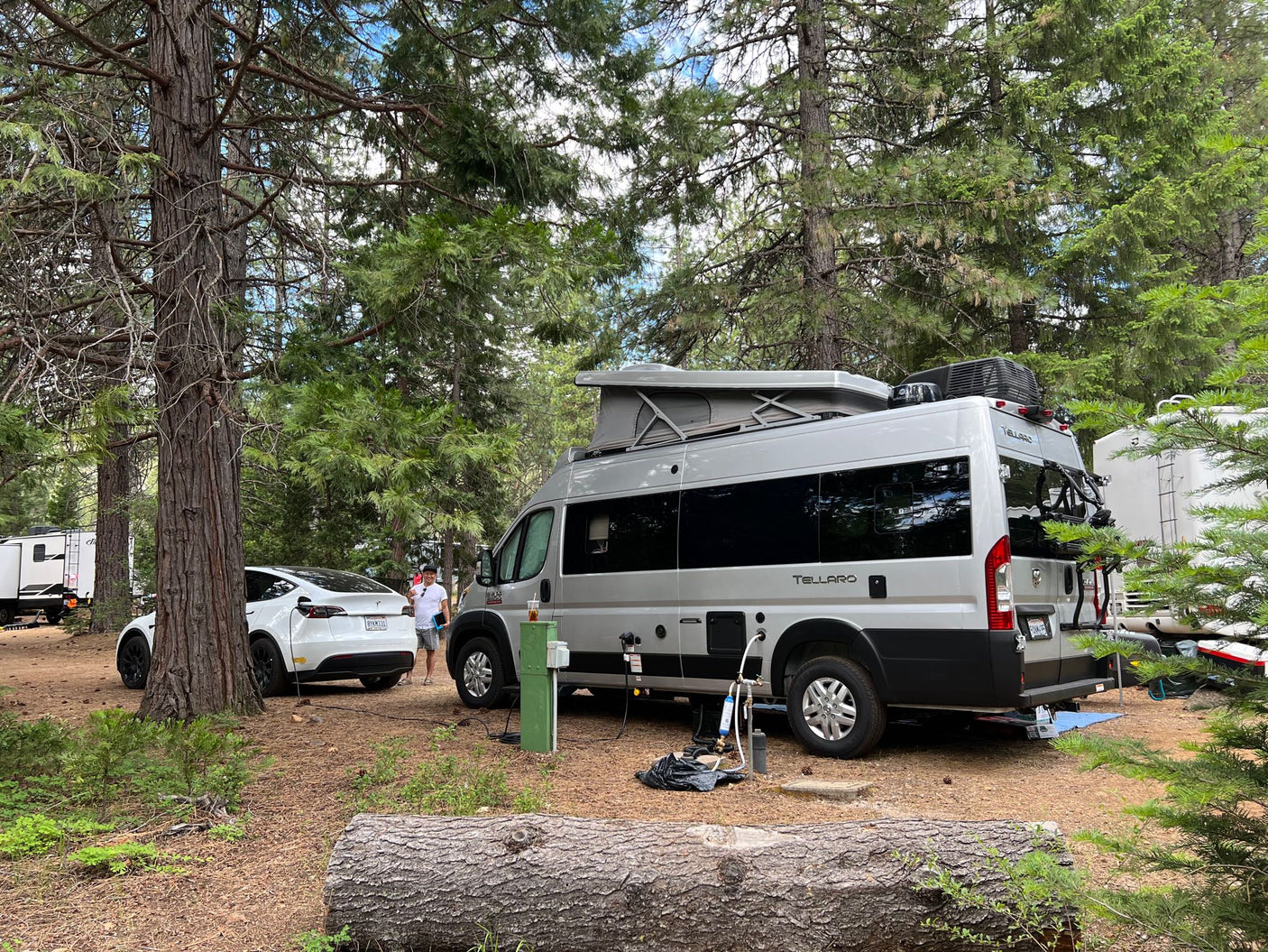A Tellaro RV rental and white Tesla Model Y charging at a treed campsite. A man stands between the vehicles.