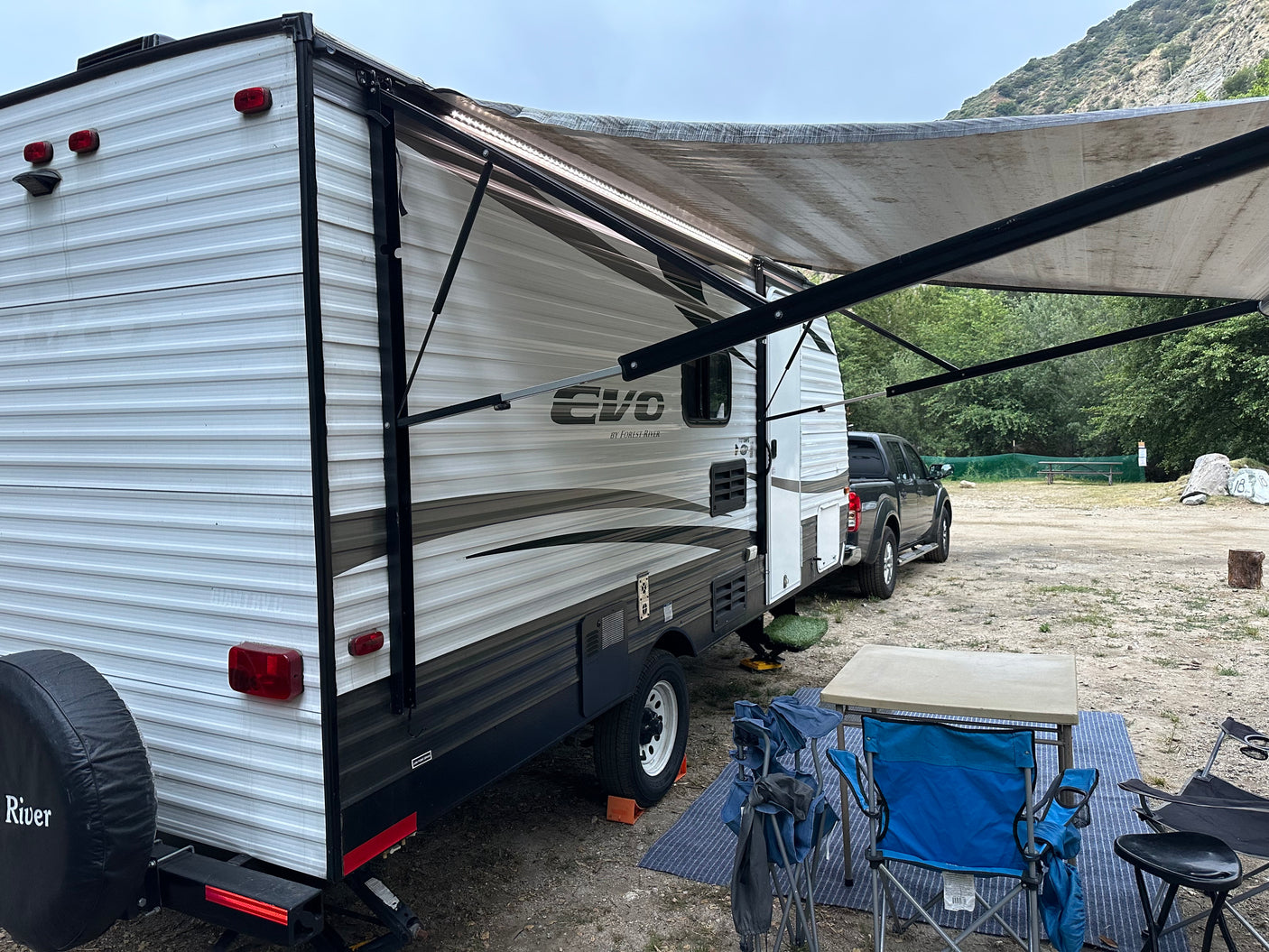 An RV rental, a white travel trailer with an extended awning, is set up at a dirt campsite with a pickup truck and chairs.