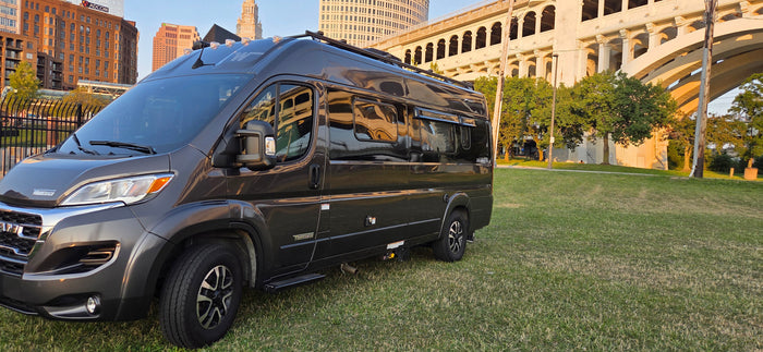 Dark gray Winnebago camper van, perfect for an rv rental, parked on green grass. City buildings & arched bridge in background.