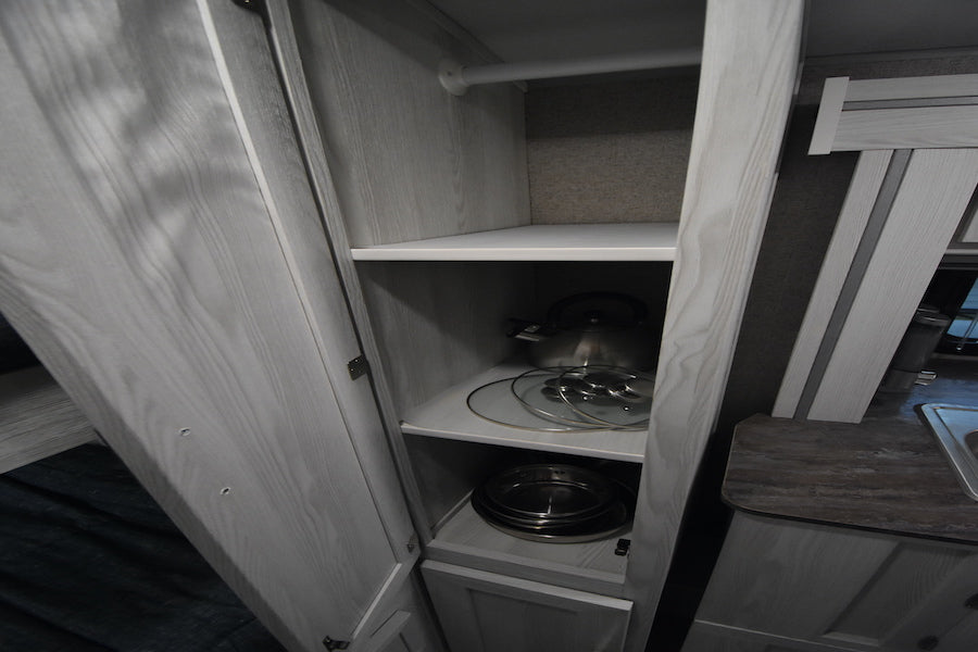 RV rental kitchen cabinet with shelves storing a kettle, pot lids, and pans, plus a hanging rod. Functional storage.
