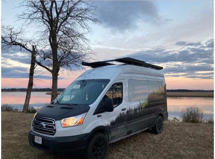 Customized Ford Transit RV rental van by a lake at dusk, with a forest graphic. Ideal for nature escapades.