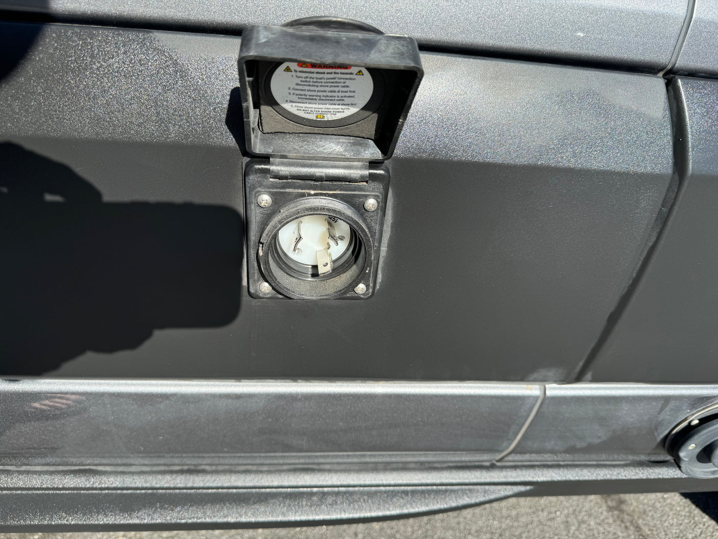 RV rental shore power inlet for external electrical hookup. The open cover displays warnings for safe cable connection/disconnection.