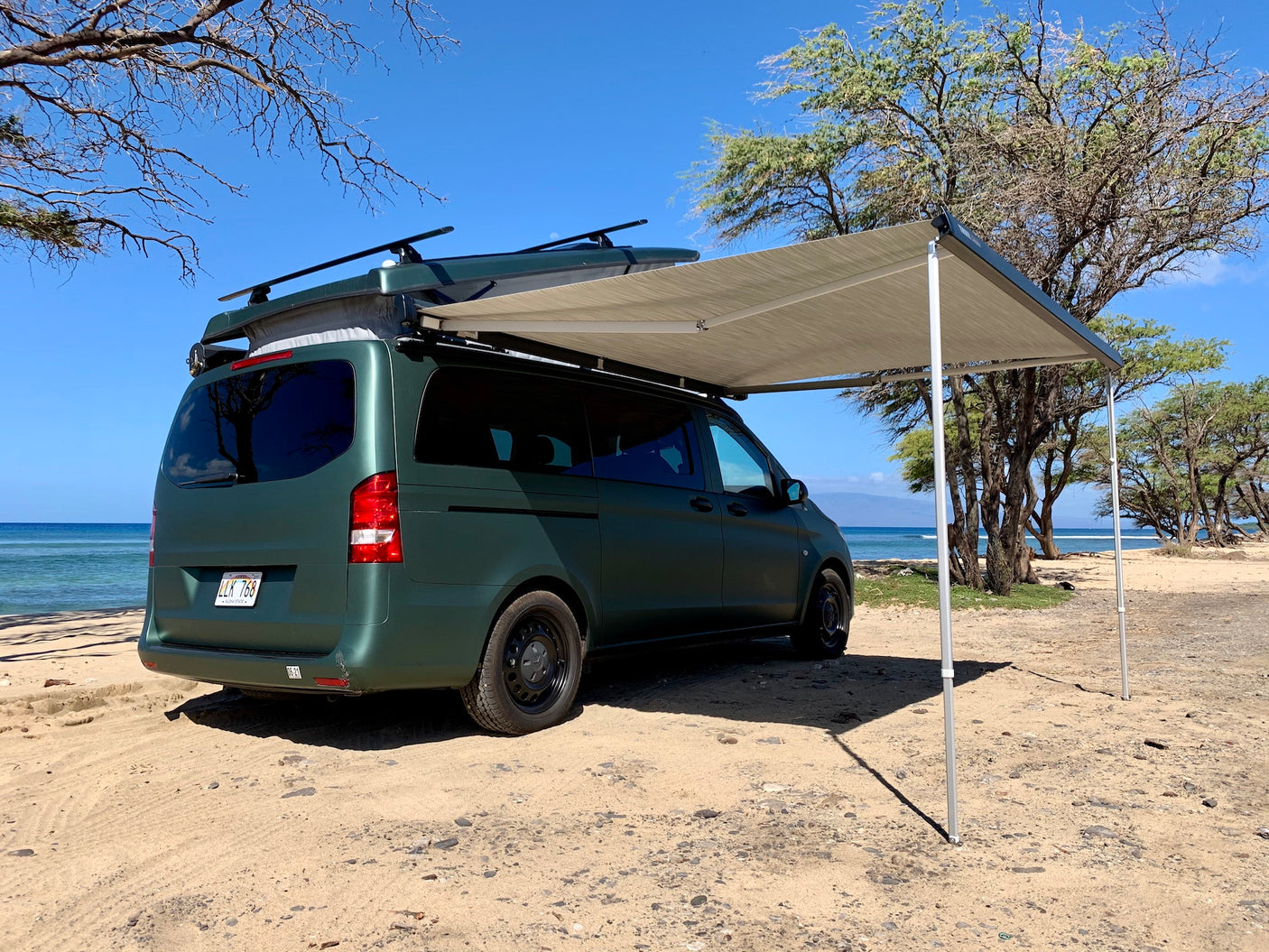 Green rv rental camper van with an awning extended, parked on a sunny Hawaiian beach.