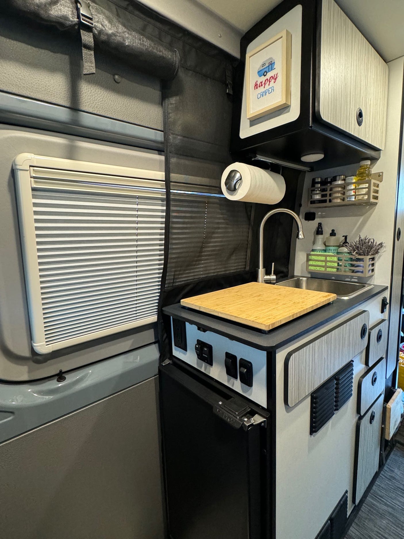 Compact RV rental interior showing a small kitchen with sink, fridge, overhead cabinets, and window blinds.