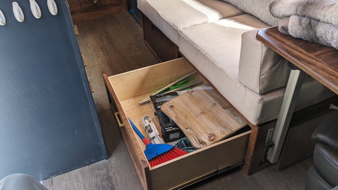 RV rental storage solution: an open drawer under a couch holds a broom, tools, and cleaning supplies, maximizing space.