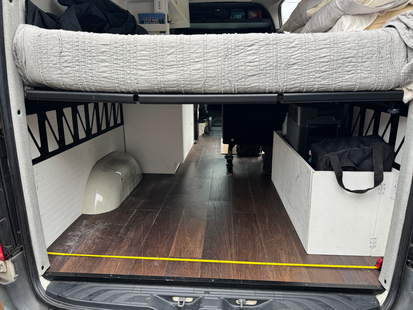 Inside a spacious RV rental camper van: elevated bed, wood floor, and integrated storage, with measuring tape on floor.