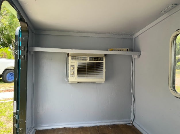 RV rental: Compact interior with a window AC unit mounted under a shelf, and clean gray walls.