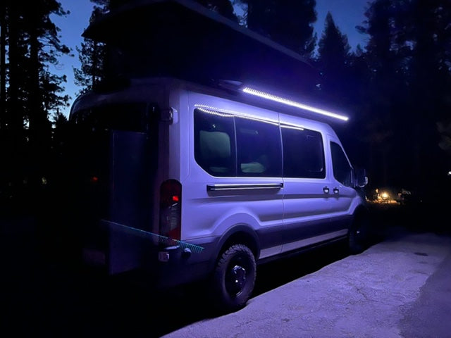 White RV rental at night, showcasing bright LED side lights & purple underglow, parked by dark trees.