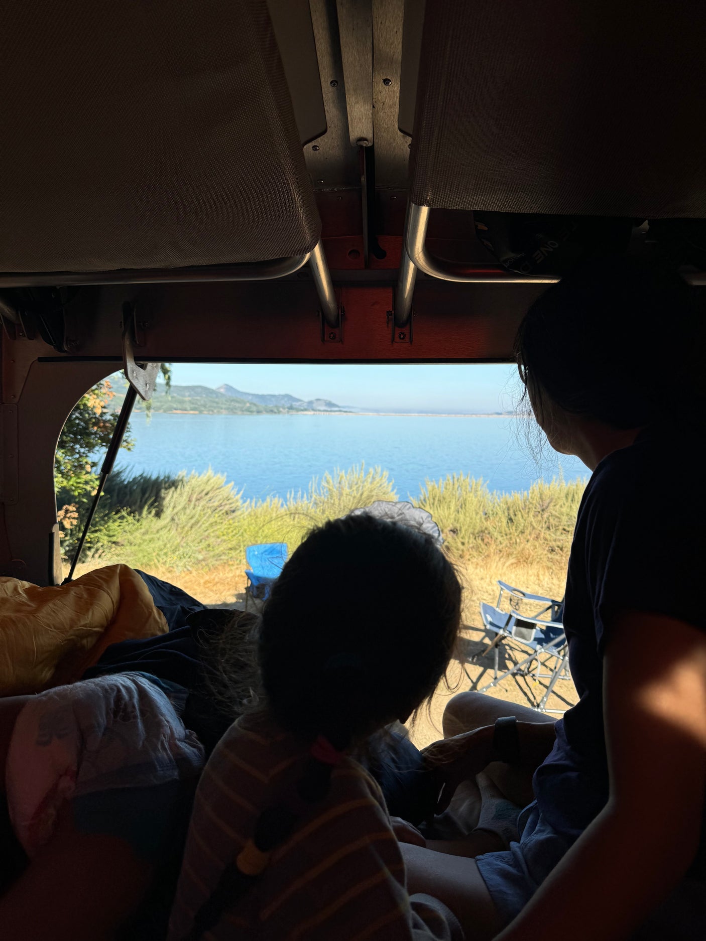 Inside our RV rental, an adult & child gaze at a serene lake & mountains. A peaceful camping view.