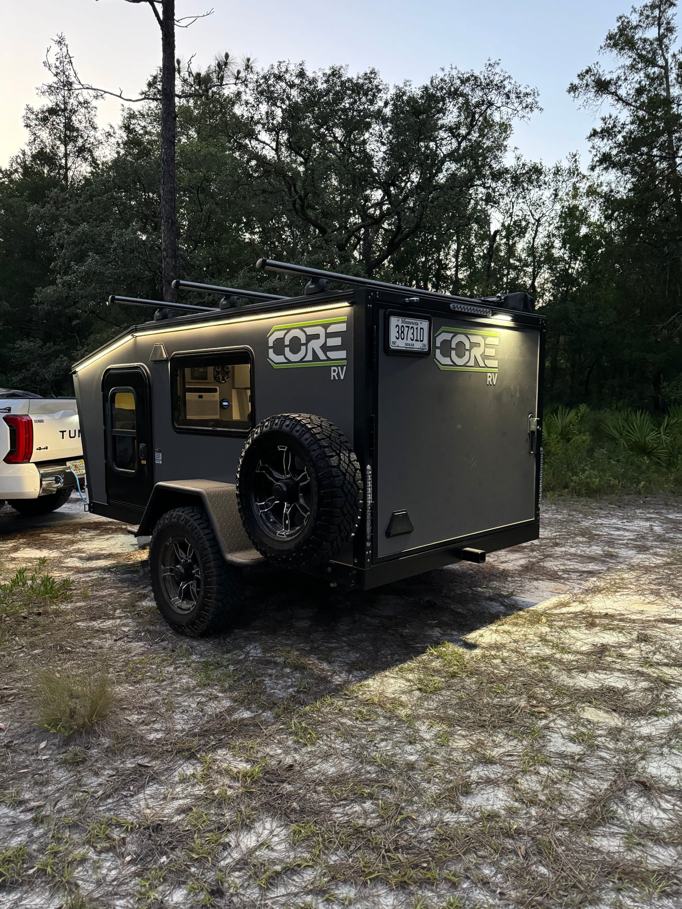 This sleek grey CORE RV rental, lit up for night, is hitched to a Tundra, set for rugged camping adventures in the forest.