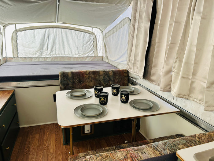 Interior of an RV rental pop-up camper with a set dining table, patterned bench seating, and a bed area.