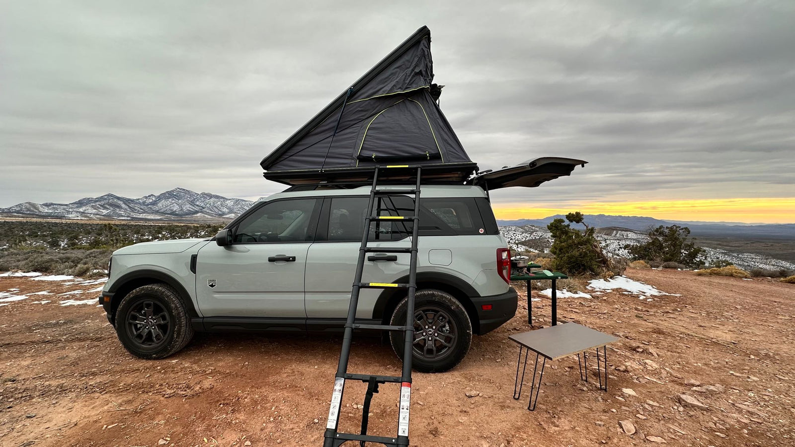 offers mountain views. A person relaxes on the bed with storage below.|Overlanding setup: Ford Bronco Sport w/ rooftop tent ready for snowy trails. An rv rental alternative for your next off-grid adventure!|Overlanding rv rental: SUV with rooftop tent set up for camping on a dirt road