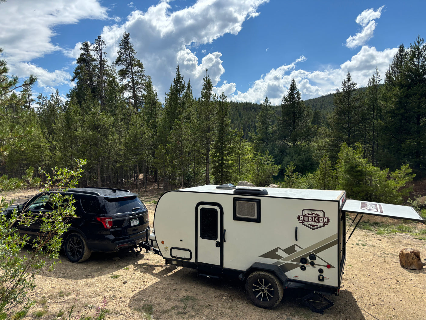 White Rubicon travel trailer (RV rental) towed by a black SUV, parked at a peaceful mountain forest campsite.